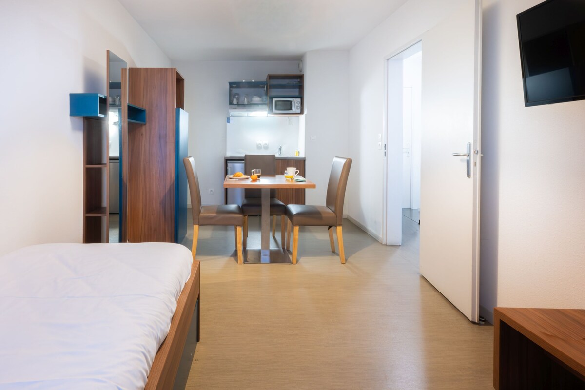 Habitación de hotel pequeña con cama, una mesa de comedor con sillas, kitchenette y televisión montada en la pared.