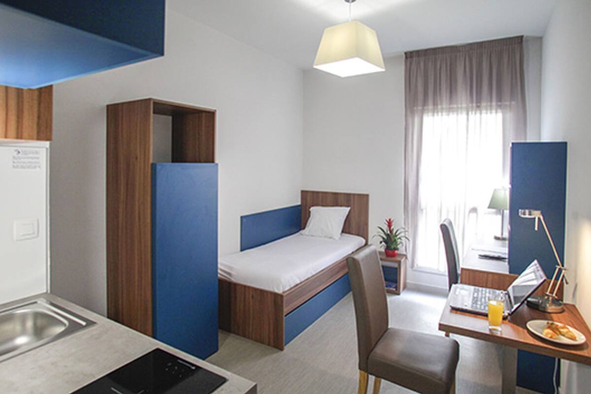 Une chambre d’hôtel de taille réduite, comprenant un lit simple, un bureau avec un ordinateur portable, une chaise, une petite table avec une assiette de nourriture et une fenêtre avec des rideaux.