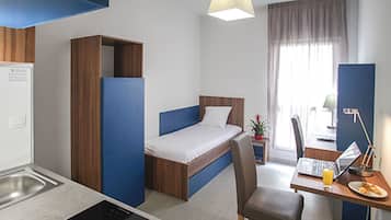 Ett kompakt hotellrum med en enkel säng, ett skrivbord med en bärbar dator, en stol, ett litet bord med en tallrik mat och ett fönster med gardiner.