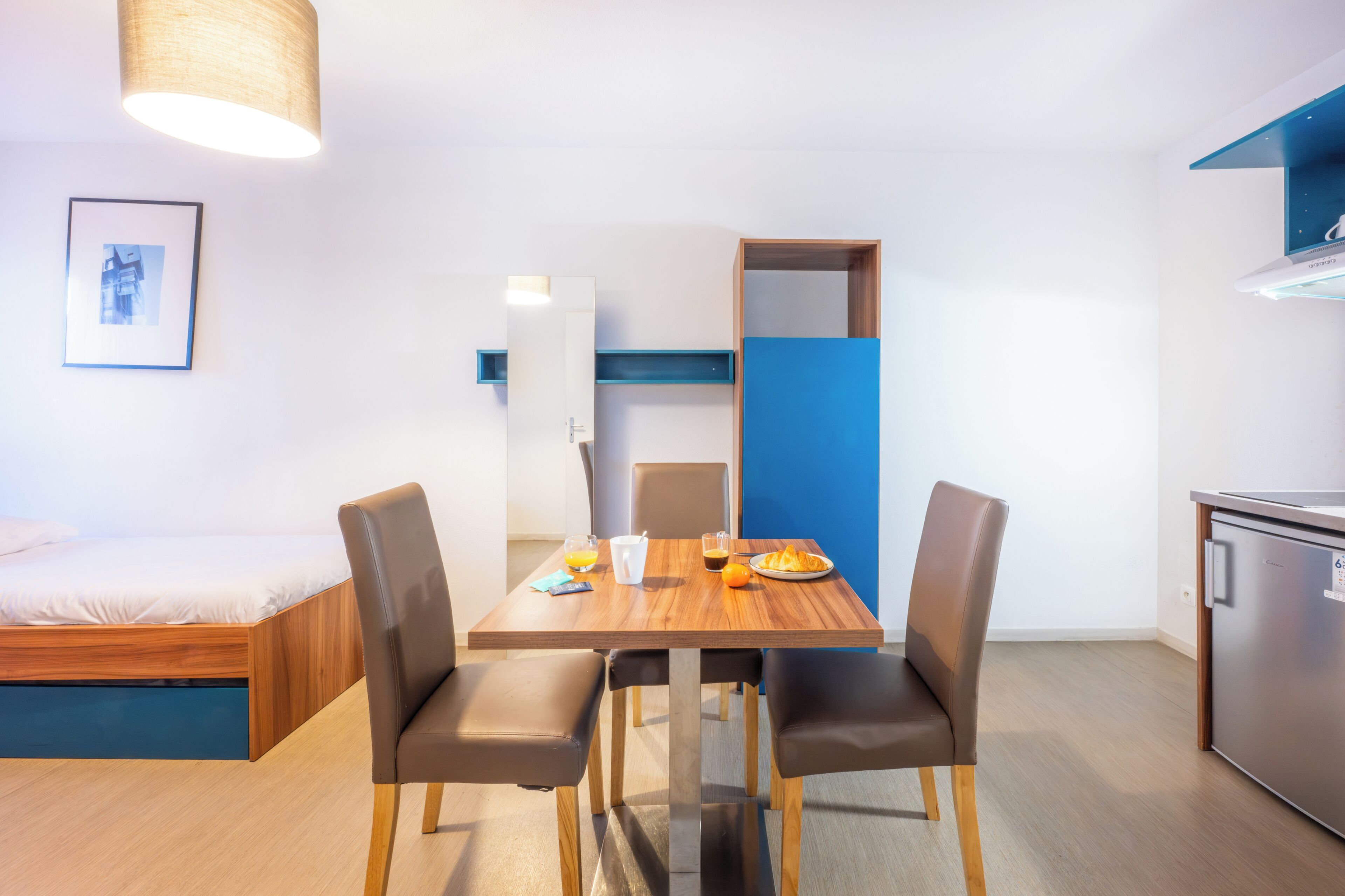 A modern room with a bed, a dining table with chairs, a small kitchen area, and a framed picture on the wall.