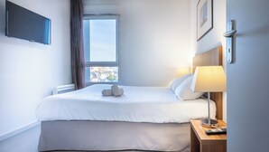 Une chambre d’hôtel avec un lit, une lampe de chevet, un téléviseur fixé au mur et une fenêtre avec des rideaux.