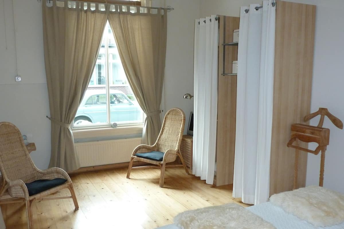 A room with a bed, two wicker chairs, a window with curtains, a radiator, and a wooden shelf.