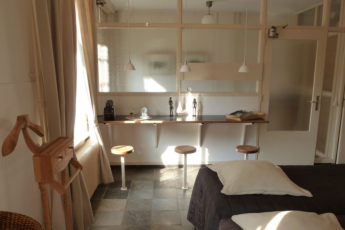A compact room with a bed, a small stool, a wooden side table, and a window with curtains.