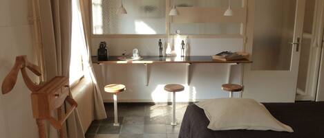 A compact room with a bed, a small stool, a wooden side table, and a window with curtains.
