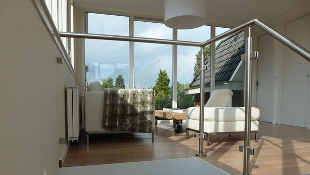 A modern living room with a large window, a white sofa, a small table, and a glass railing. - Hotel Het Oude Postkantoor (Brummen)