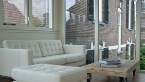 A modern living room with a white sofa and ottoman, a wooden coffee table with books, and large windows overlooking a brick building. - Hotel Het Oude Postkantoor (Brummen)