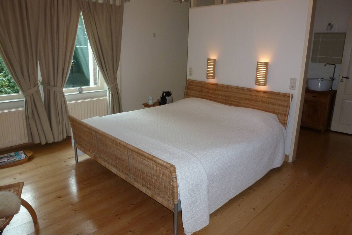 A bedroom with a wooden bed, beige curtains, a nightstand, and a wall-mounted light fixture.