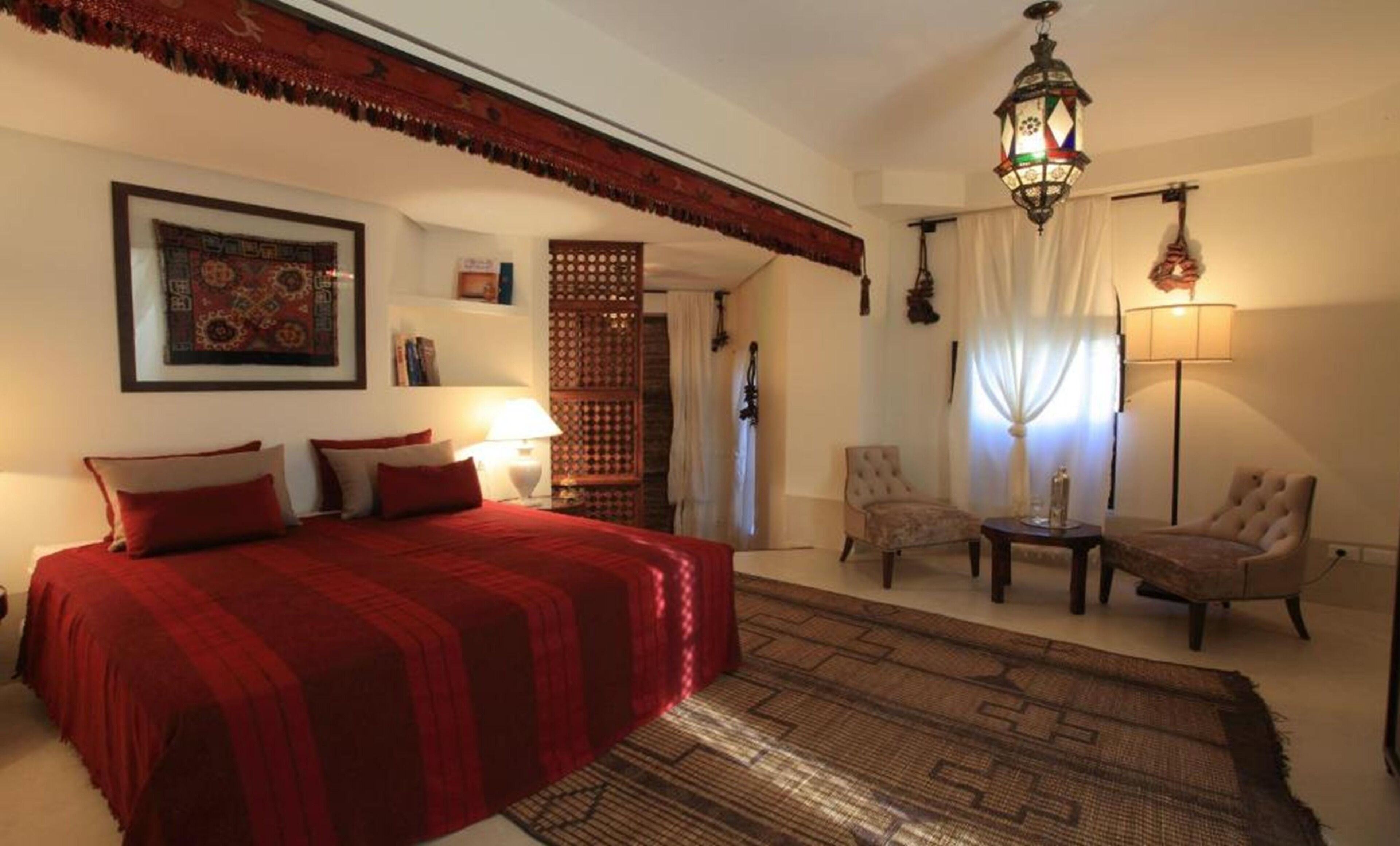 A bedroom with a large bed, two chairs, a small table, a decorative wall hanging, and a hanging lamp.