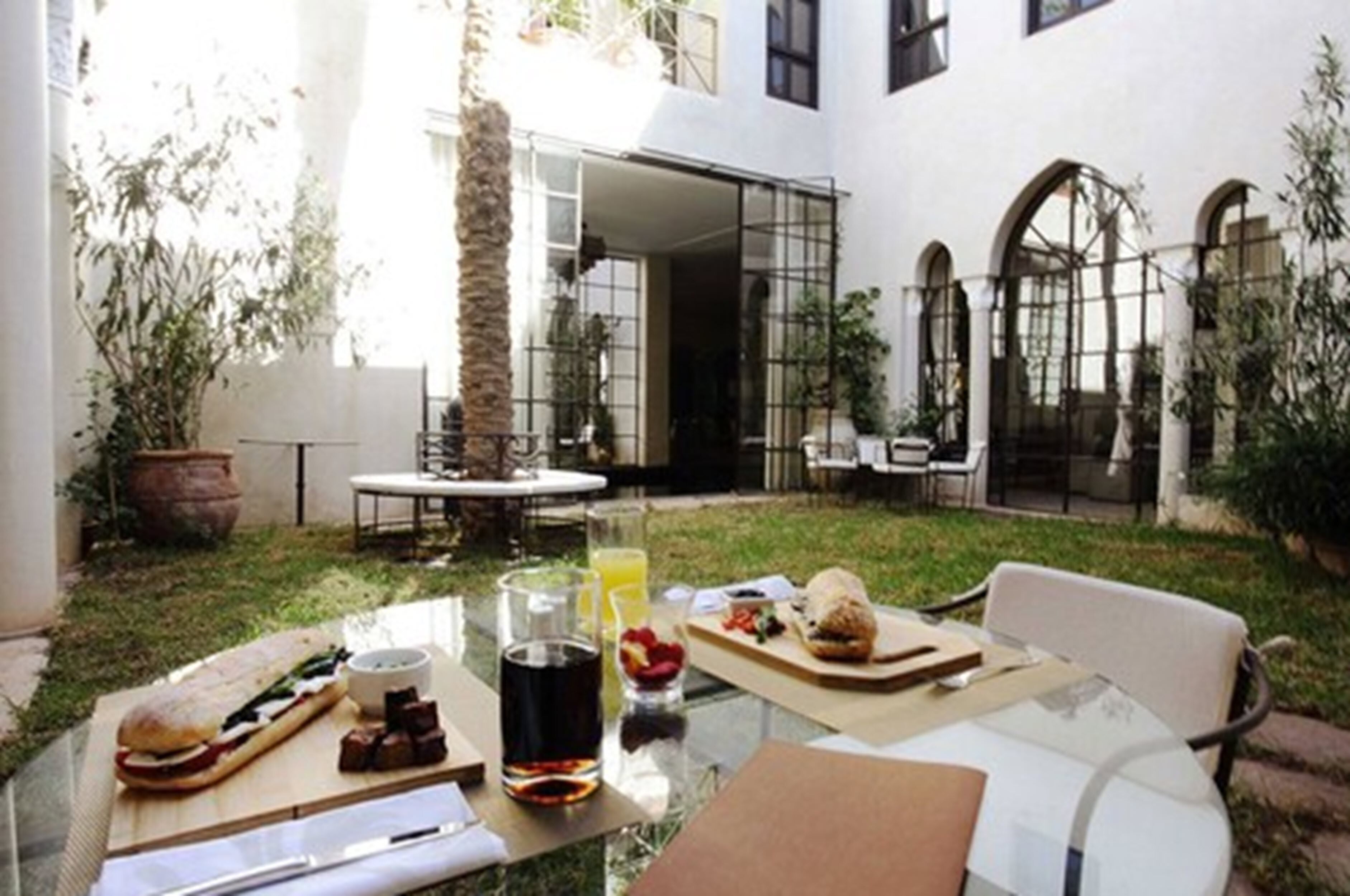 Una mesa de vidrio con comida y bebidas, un edificio blanco con ventanas arqueadas y un área verde al aire libre.