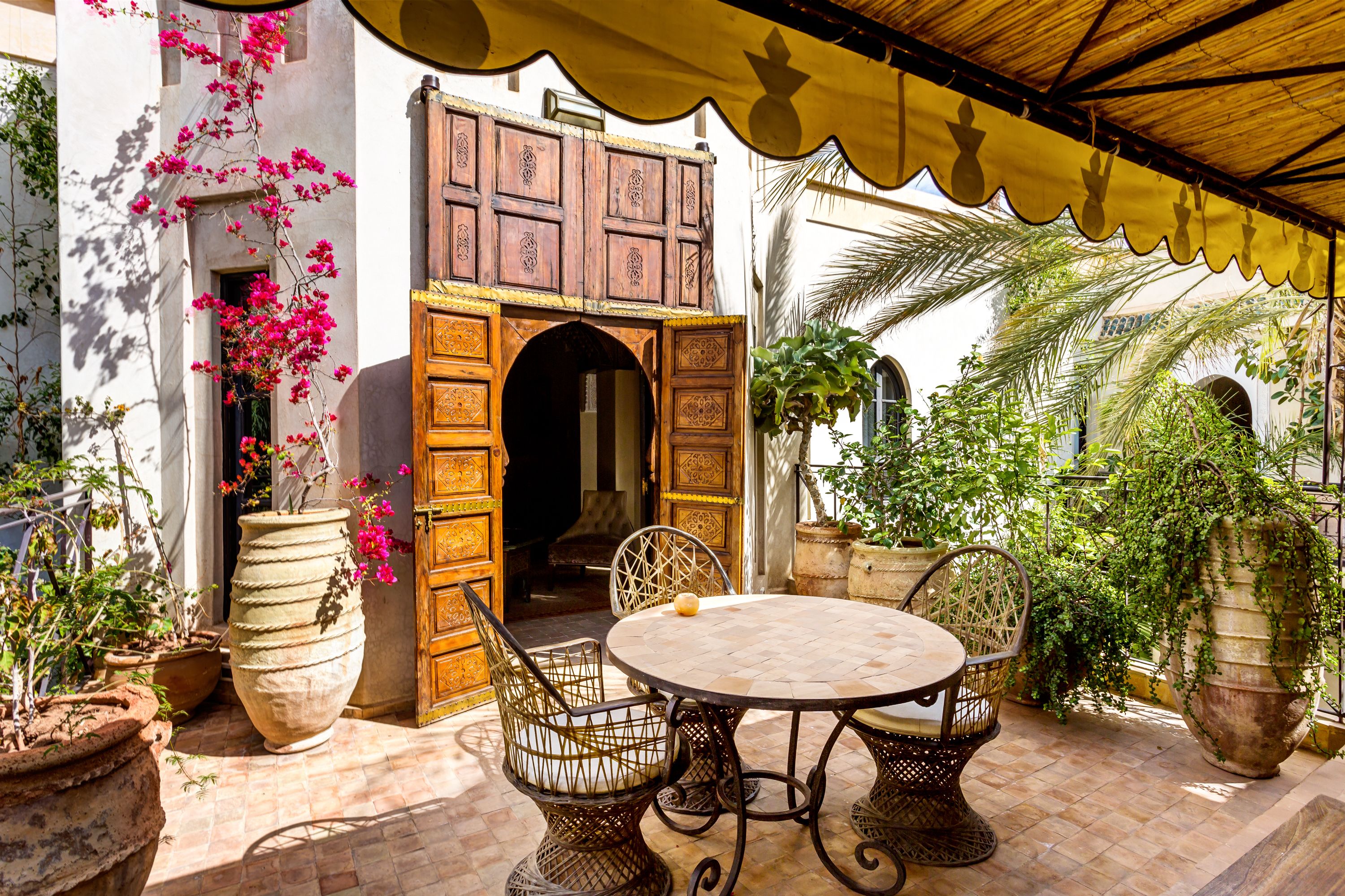 An outdoor patio with a round table and wicker chairs, surrounded by large potted plants and a wooden door with intricate carvings.