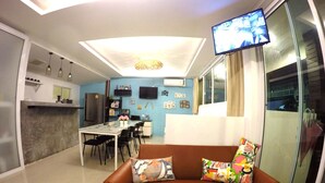 Una sala moderna con un sofá de cuero marrón, una mesa de comedor con sillas, un área de cocina con un refrigerador y un televisor de pantalla plana montado en la pared.