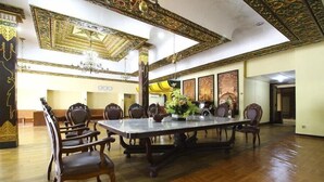 Een ruime kamer met een grote eettafel, omringd door stoelen, decoratieve plafondornamenten en kunstwerken in lijsten aan de wanden.