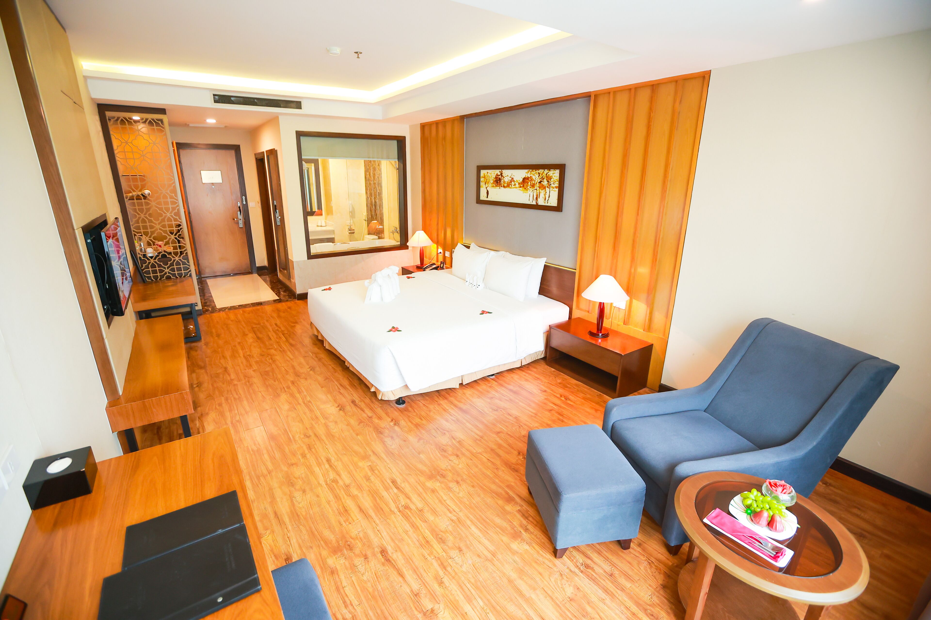 Ein Hotelzimmer mit einem großen Bett, einem blauen Sessel, einem kleinen Tisch mit Obstschale und einem Holztisch.