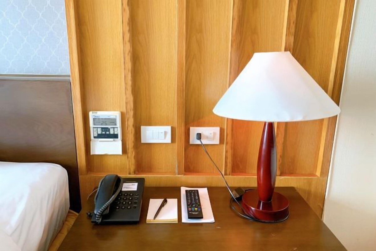 Habitación de hotel con cabecera de madera, mesita de noche con lámpara, teléfono, control remoto y toma de corriente.