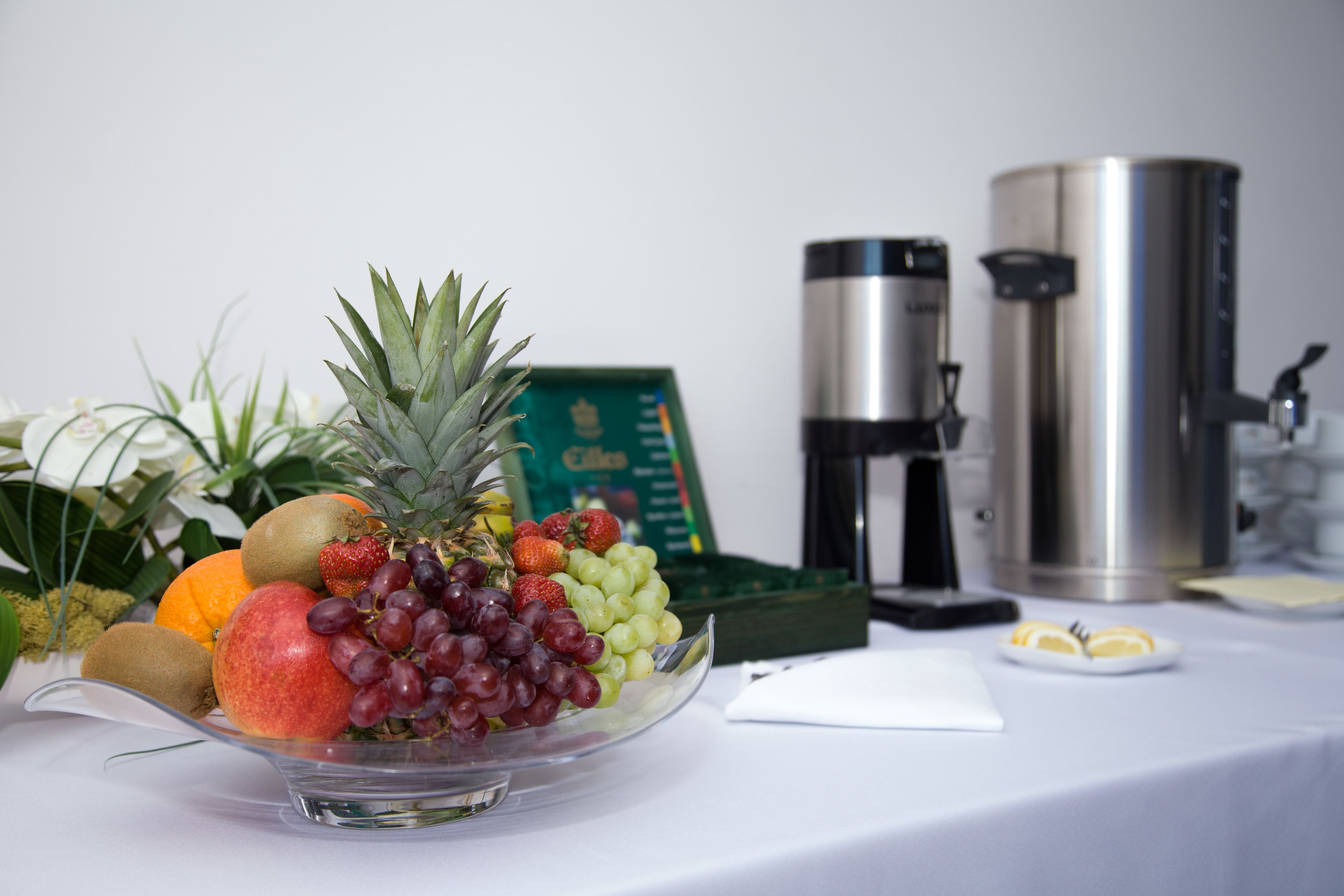 A fruit bowl on a table with a coffee maker and a thermos in the background.