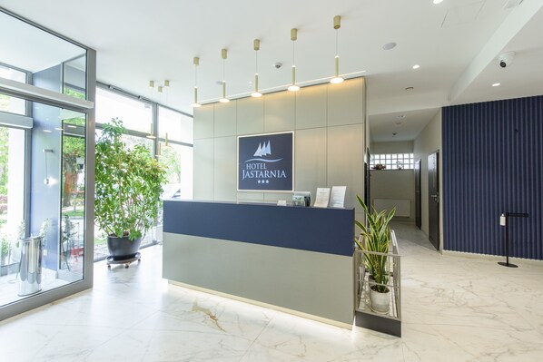 A modern hotel reception desk with a logo, potted plants, and a view of the outdoors. - Hotel Jastarnia (Jastarnia)