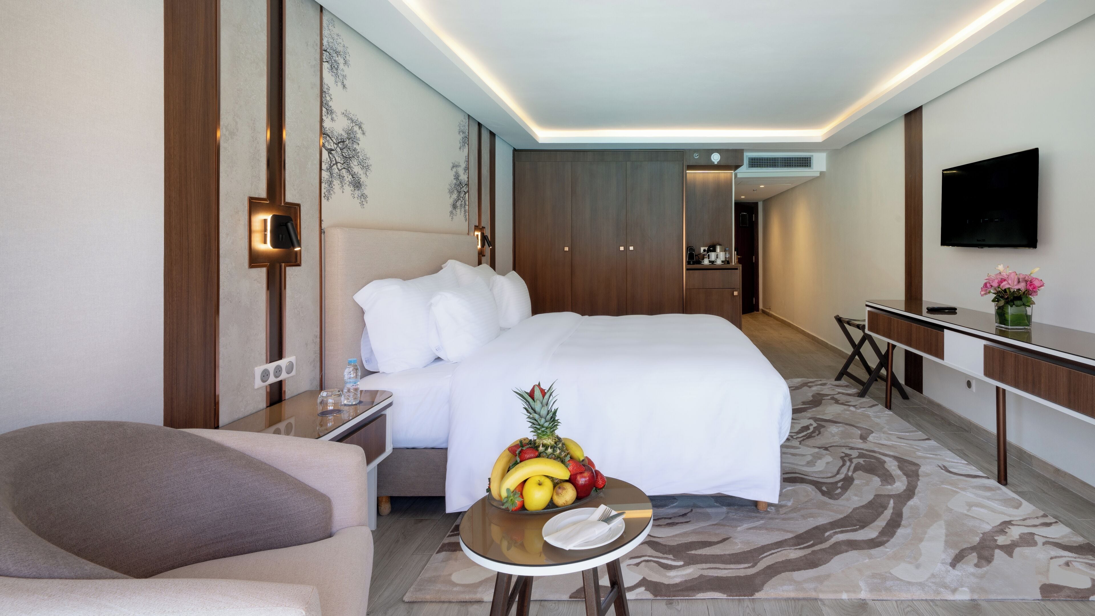 A hotel room with a bed, bedside table, a small round table with fruit, a sofa, a desk, a TV, and a wall-mounted air conditioner.