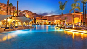 Una amplia piscina interior con sillones de descanso y palmeras, rodeada por un hotel con ventanas arqueadas e iluminación cálida.