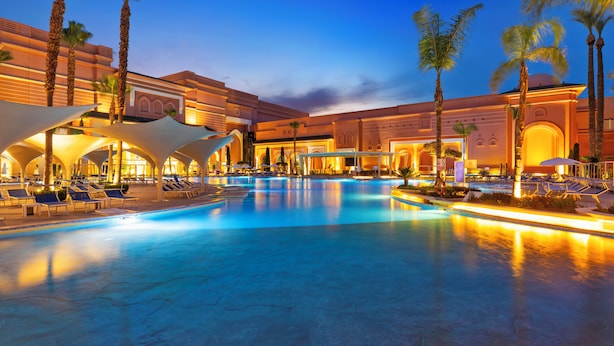 Una amplia piscina interior con sillones de descanso y palmeras, rodeada por un hotel con ventanas arqueadas e iluminación cálida.
