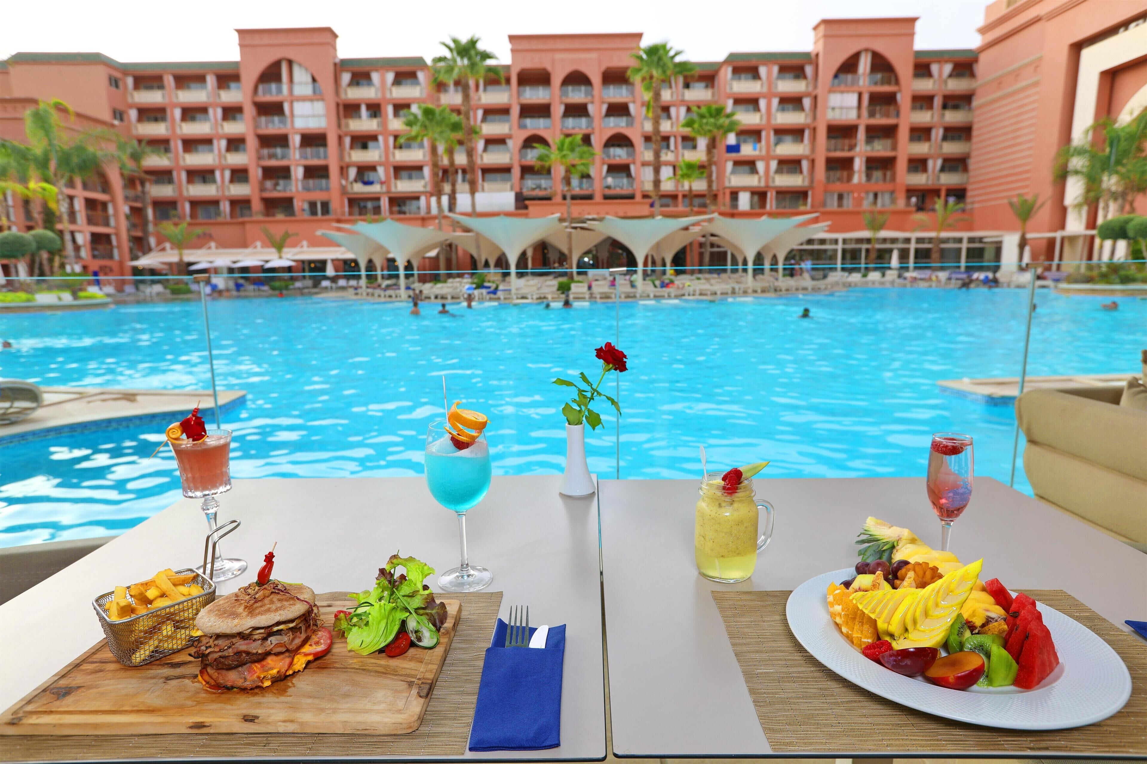 Una mesa de comedor con comida y bebidas junto a una piscina, con vistas a un hotel con piscina y palmeras.