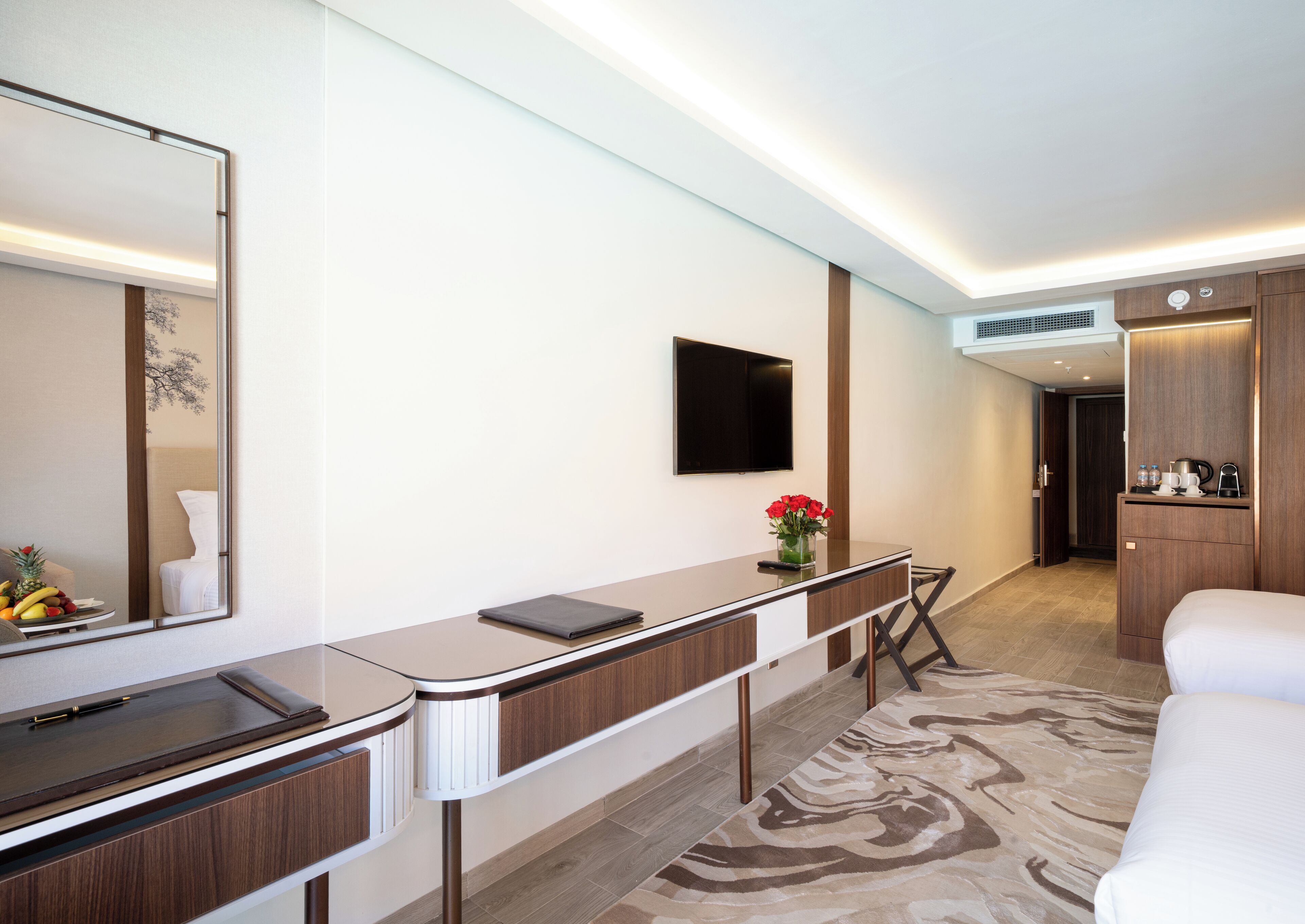 A modern hotel room with a large mirror, a flat-screen TV, a desk with a laptop, a bed, and a small table with a vase of flowers.