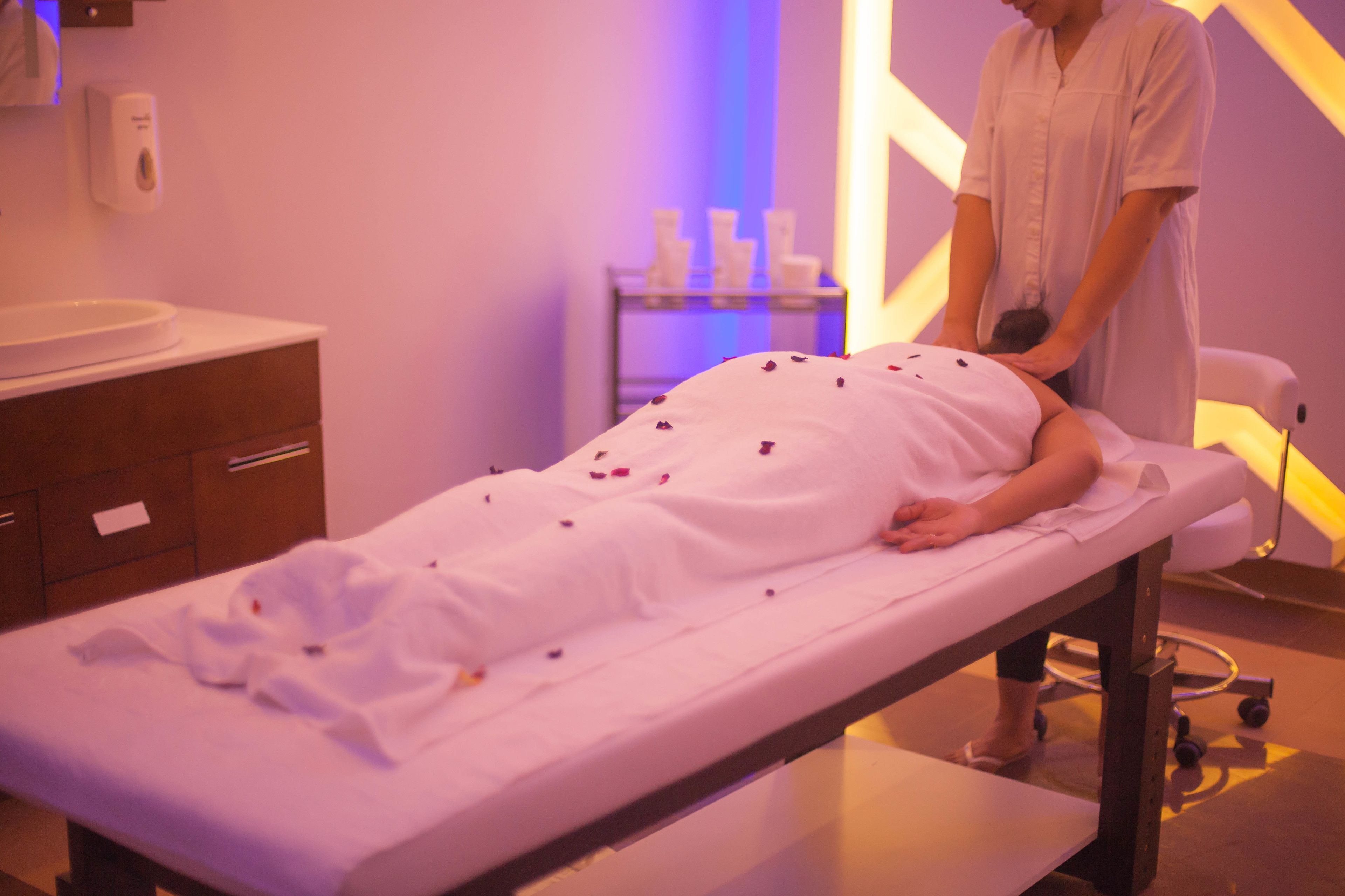 Une salle de massage avec une table de massage, une personne recevant un massage et une étagère avec des bouteilles en arrière-plan.