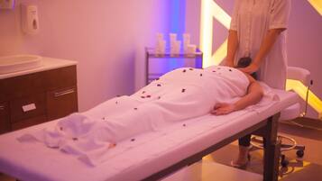 A massage therapy room with a massage table, a person receiving a massage, and a shelf with bottles in the background.