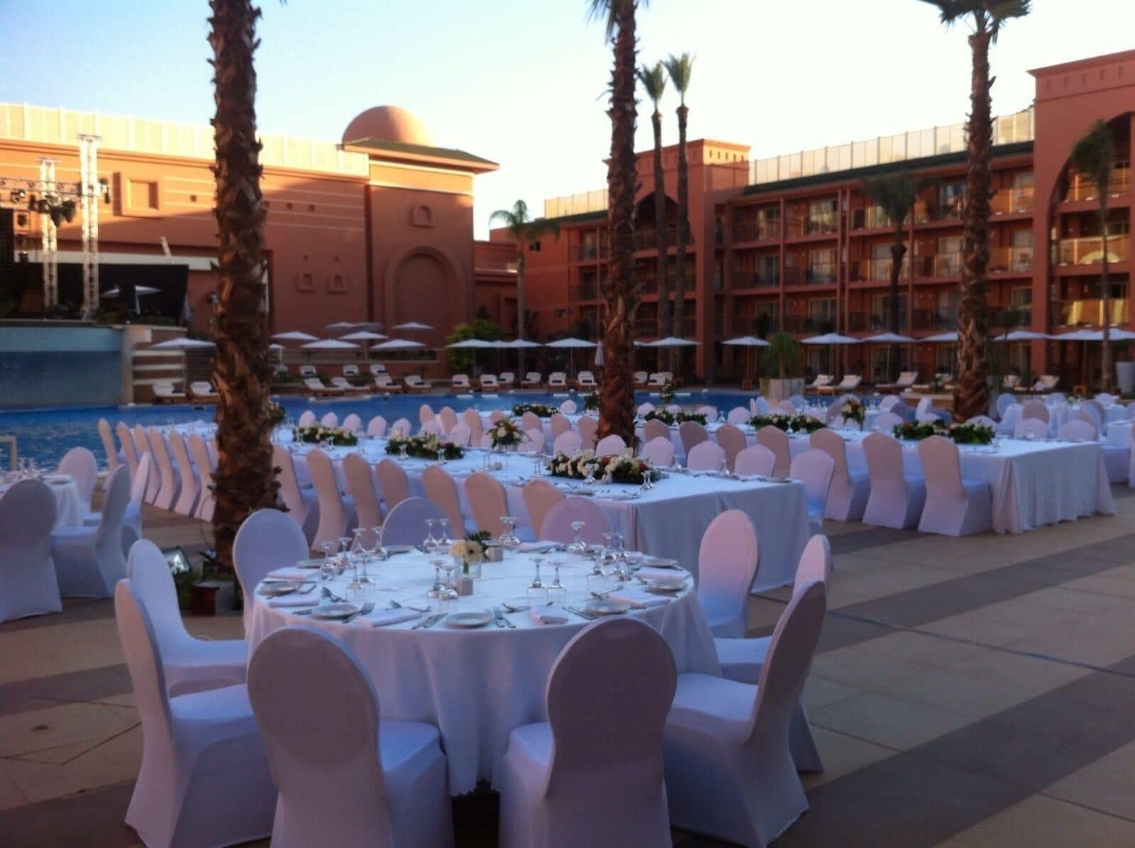 An outdoor banquet setup with round tables, white chairs, and a pool in the background.