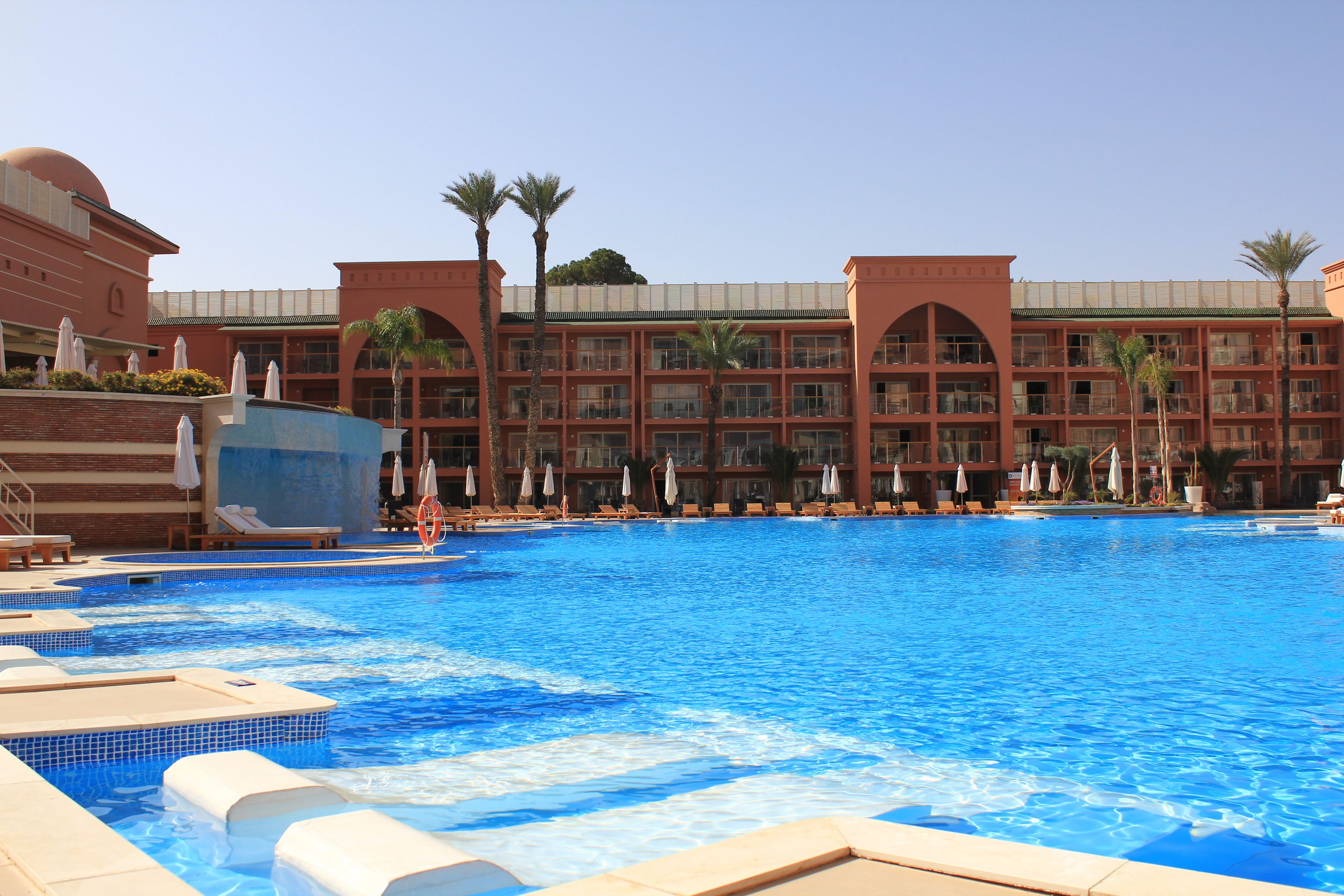 Una gran piscina con sillas de descanso y sombrillas, rodeada por un edificio de hotel de varios pisos con balcones y palmeras.