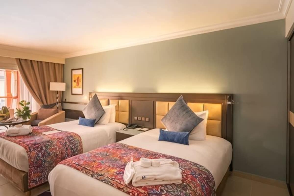 A hotel room with two beds, a nightstand, a lamp, and a window with curtains.