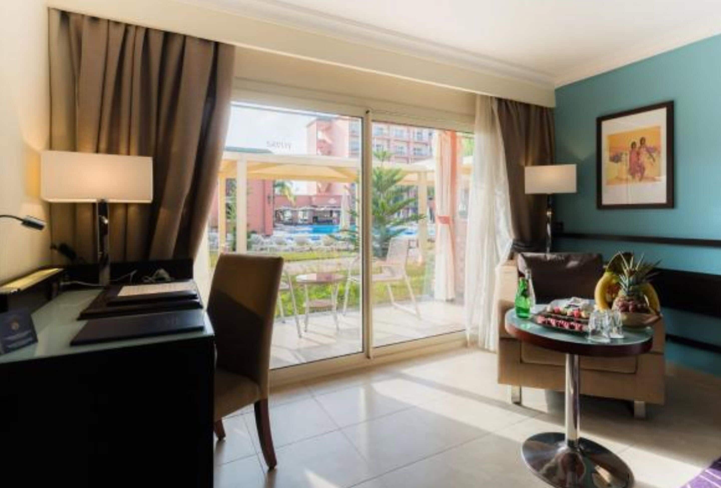 Habitación de hotel con escritorio, silla y una mesa con fruta y una bebida. Se ve una piscina y edificios desde el exterior.