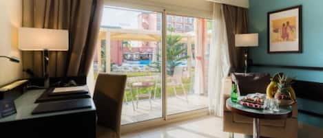 Habitación de hotel con escritorio, silla y una mesa con fruta y una bebida. Se ve una piscina y edificios desde el exterior.