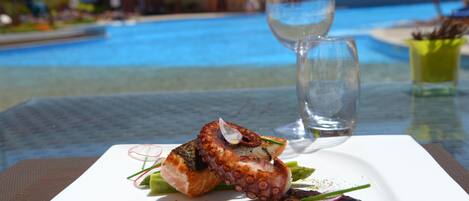 Una mesa de comedor con un plato de pulpo a la parrilla y verduras, un vaso de agua, y una piscina al fondo.