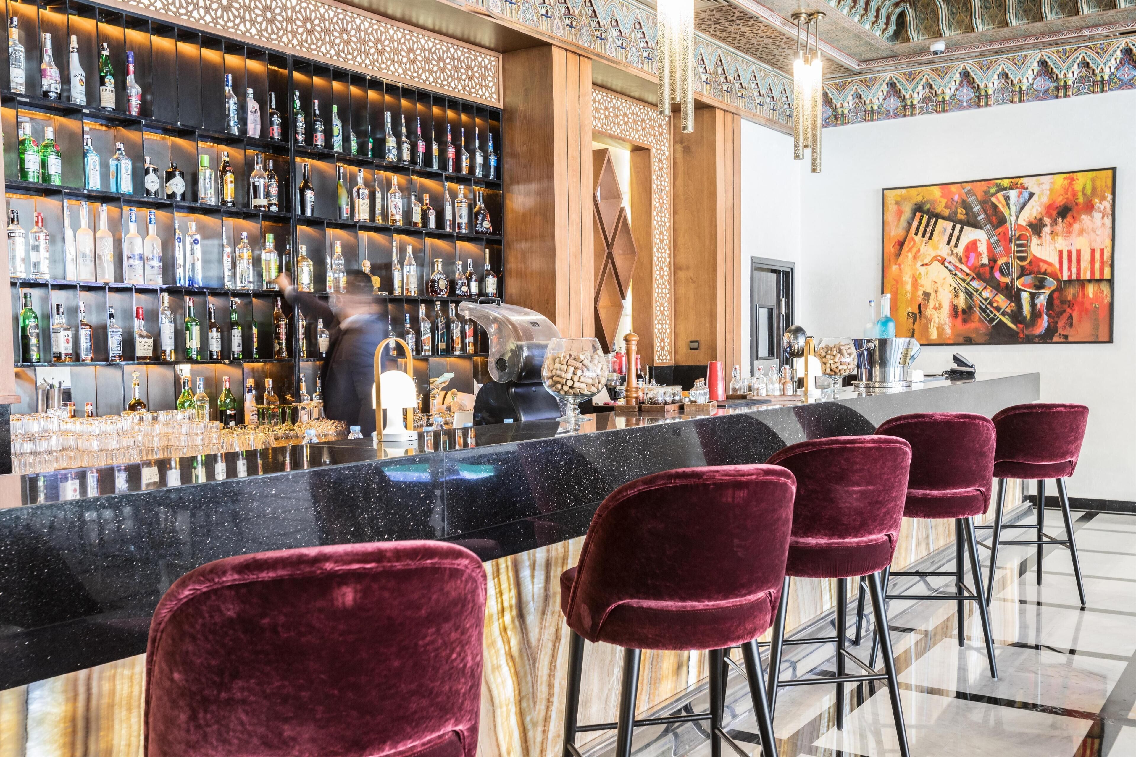 En bar med en lang bardisk, flere hylder med spiritusflasker og røde fløjelsbarstole.