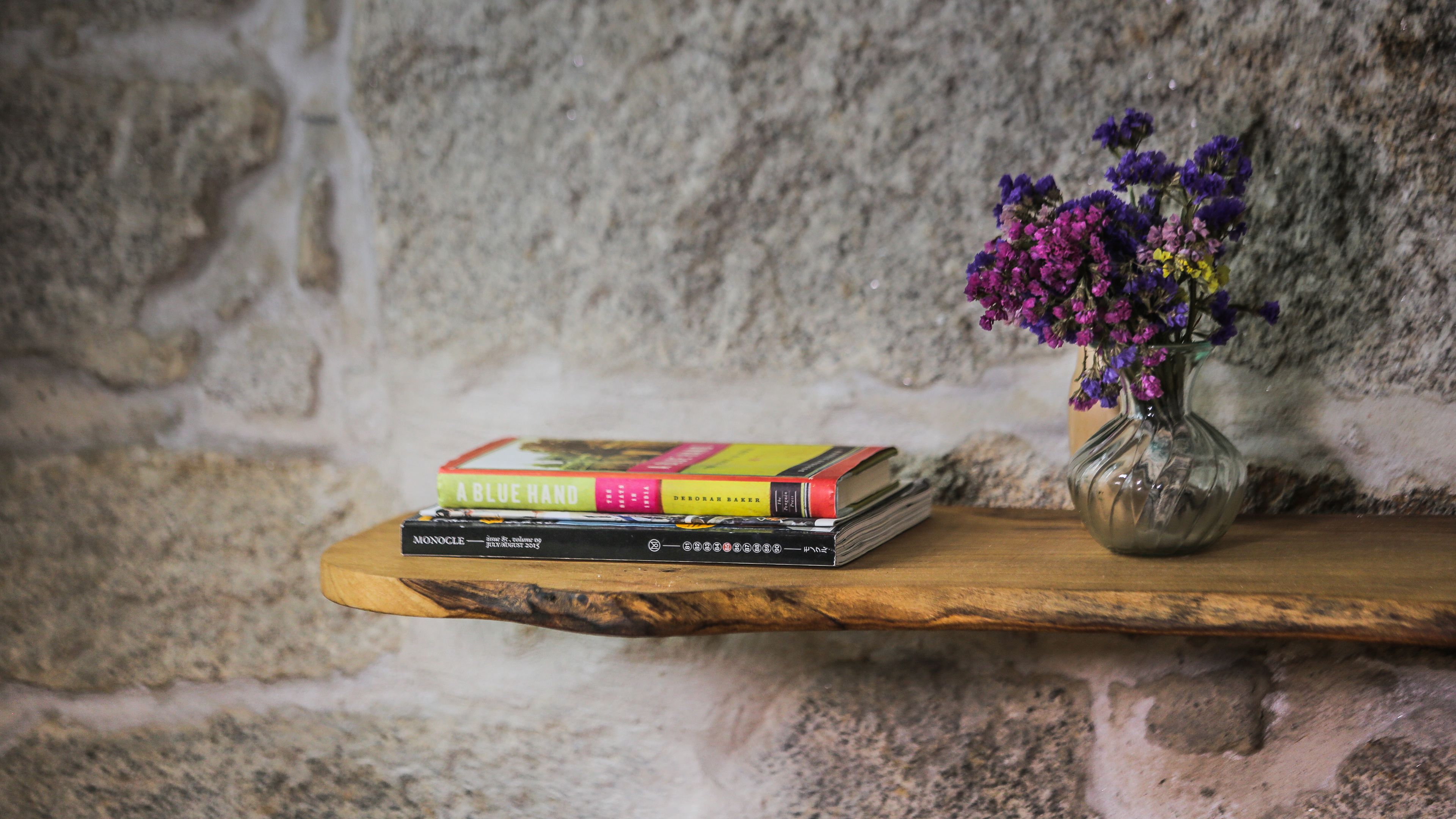 Une étagère en bois sur laquelle sont empilés des livres, et un vase contenant des fleurs violettes, le tout adossé à un mur de pierre.