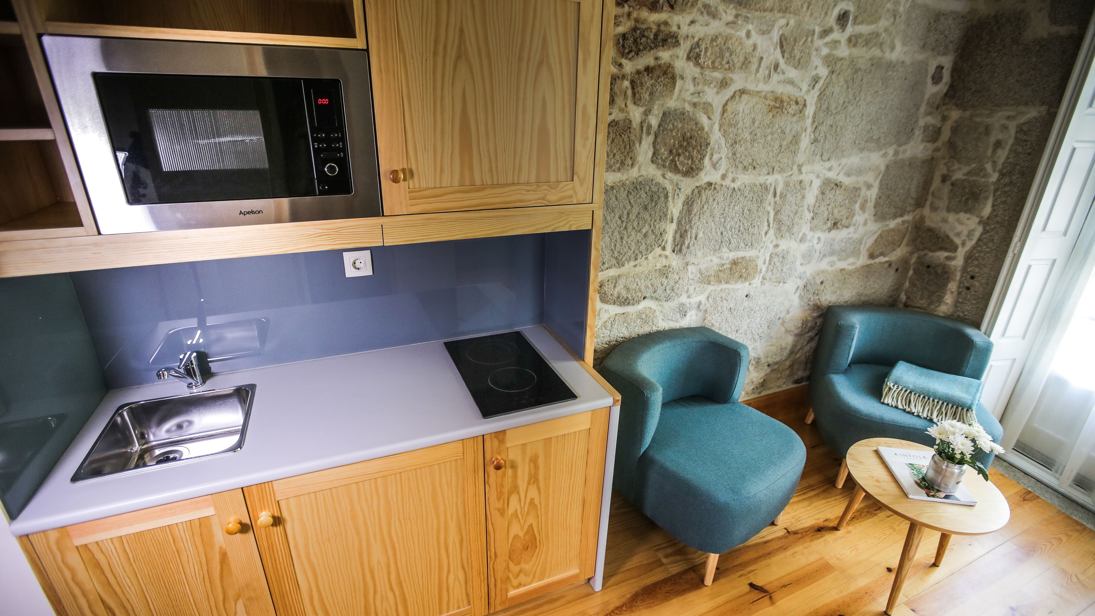 Une cuisine compacte équipée d’un four à micro-ondes, d’un évier et de placards en bois. On y trouve deux chaises bleues et une petite table sur laquelle est posé un vase de fleurs.