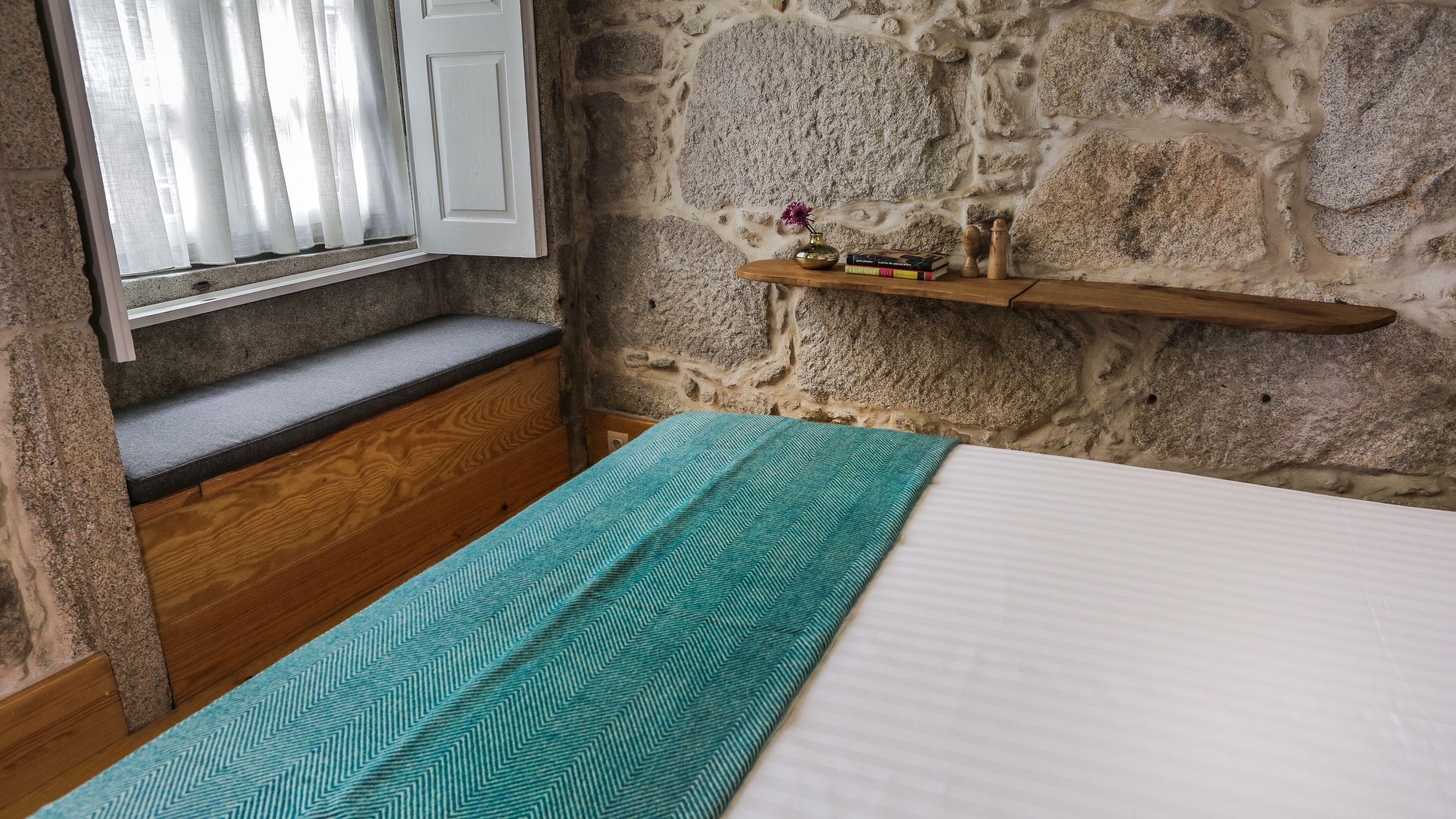 A bedroom with a stone wall, a wooden bench, a shelf with books and a vase, and a bed with a patterned blanket.