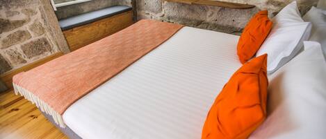 Una cama con ropa de cama blanca y naranja, un plaid estampado y un banco de madera junto a la ventana.