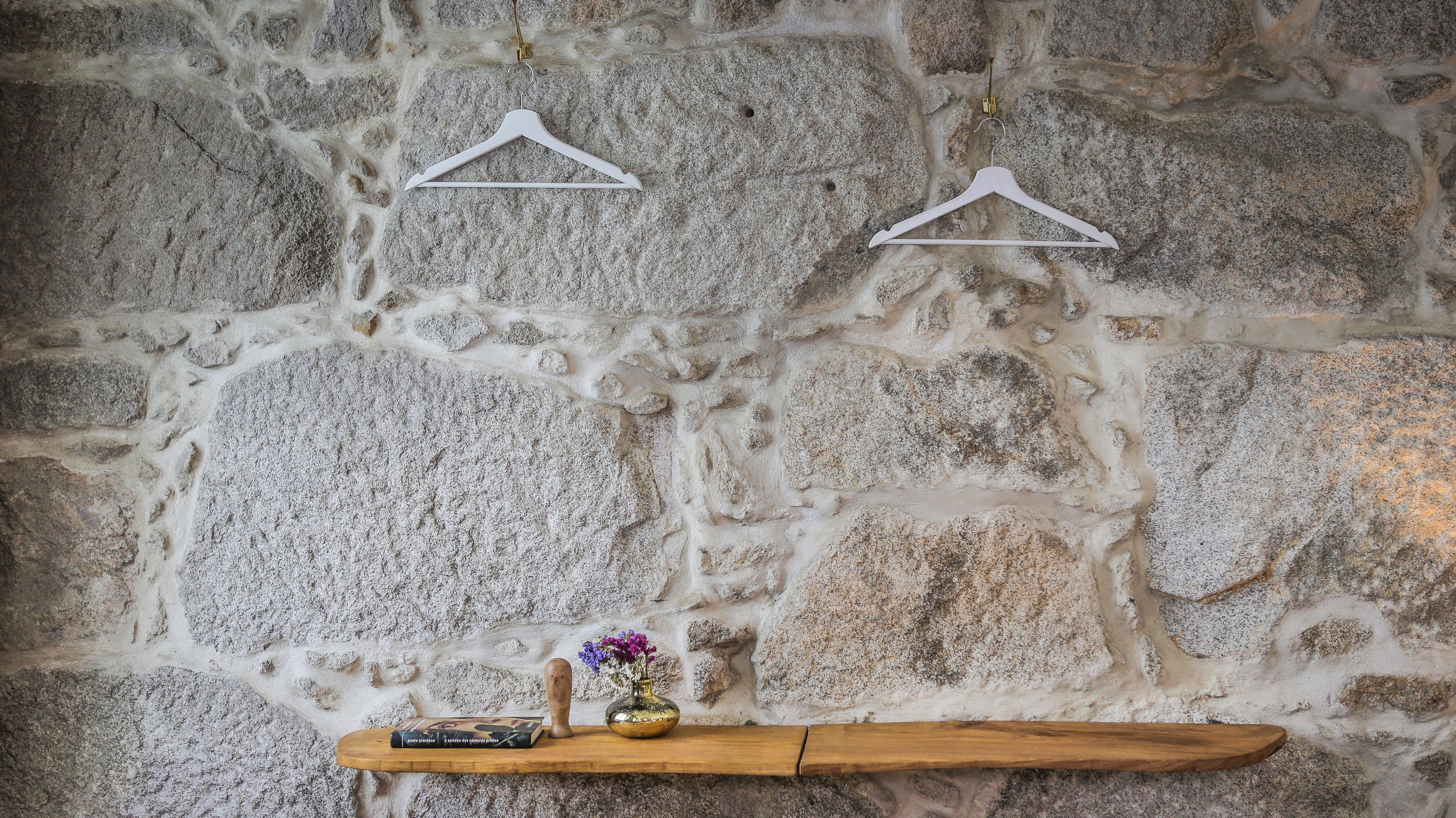 A stone wall with two white hangers and a wooden shelf with a book and a vase of flowers.