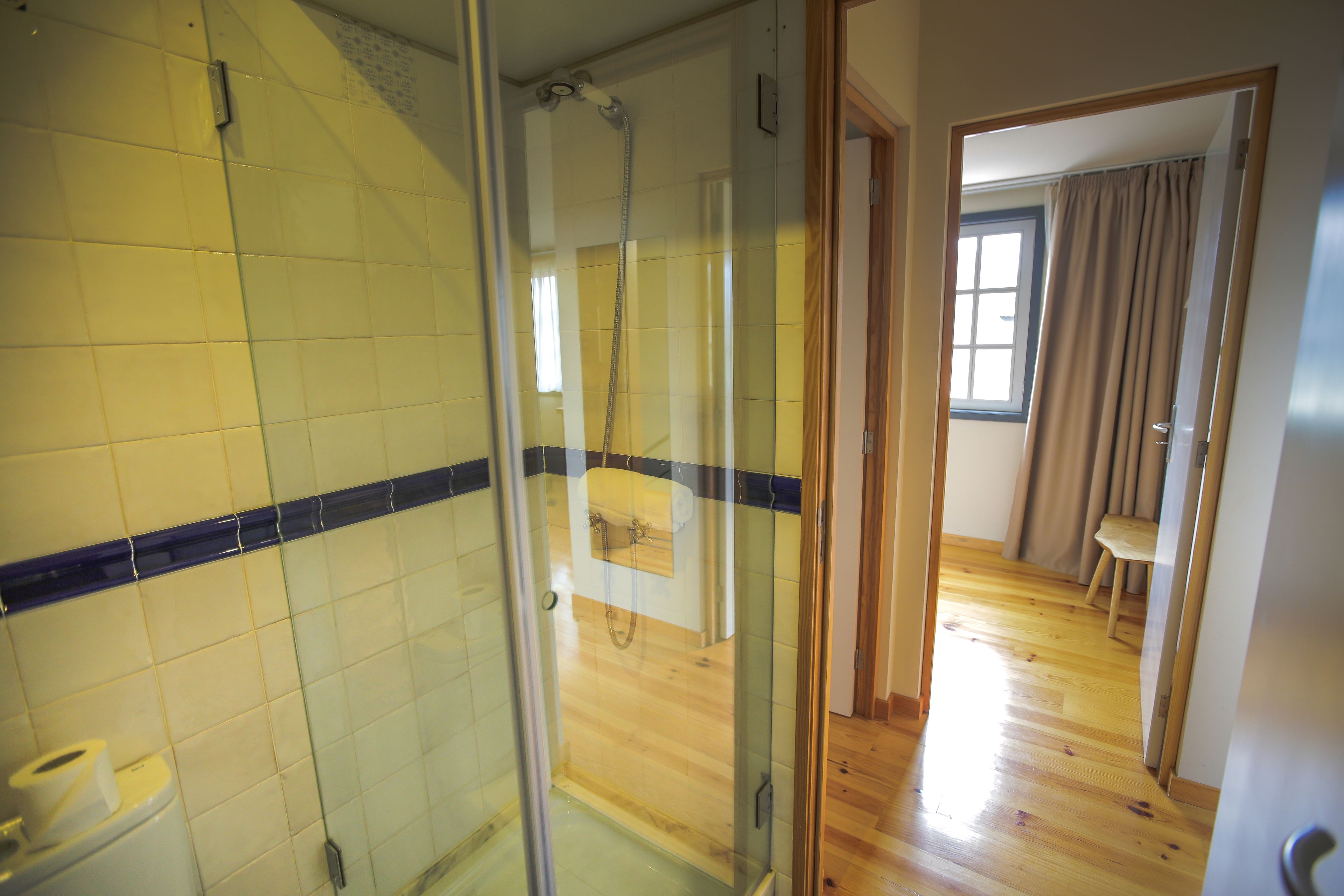 A bathroom with a glass shower enclosure, a wooden floor, and a window with curtains.