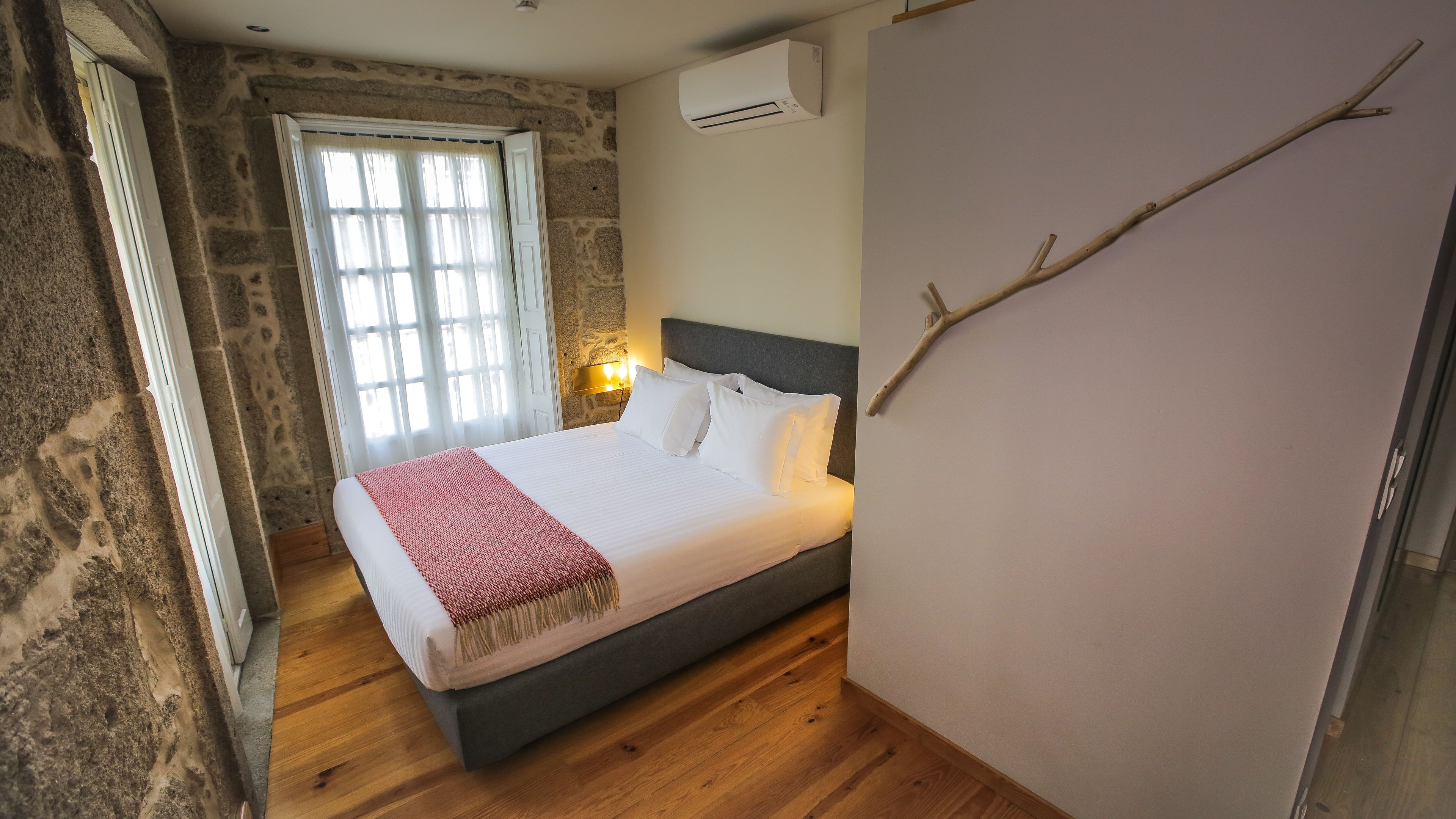 Une chambre avec un mur en pierre, un lit avec une literie blanche et un plaid rouge, une fenêtre avec des rideaux et un climatiseur fixé au mur.