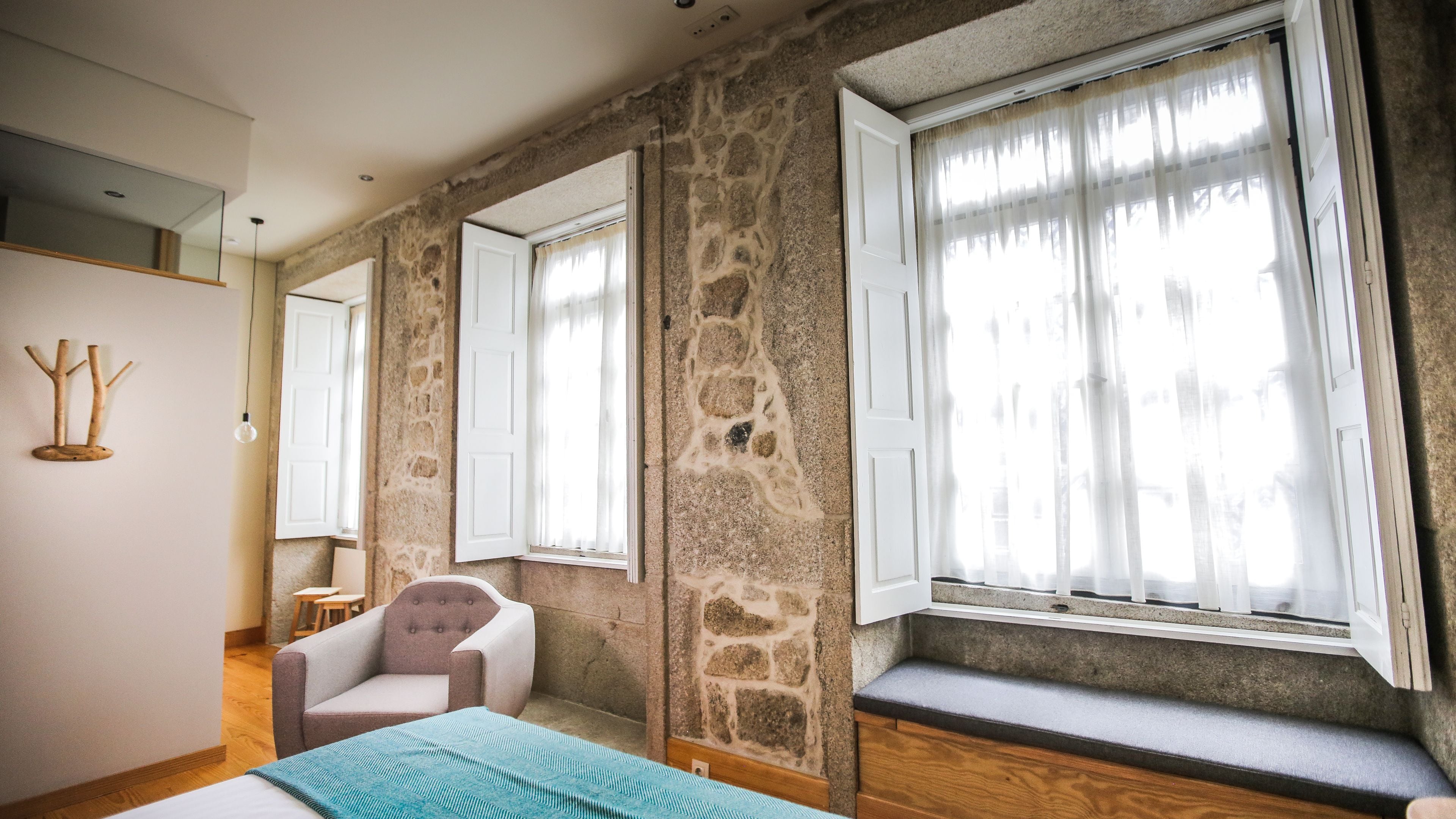 A room with stone walls, a bed, a chair, and large windows with sheer curtains.