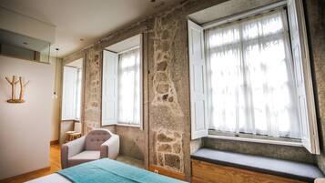 Una habitación con paredes de piedra, una cama, una silla y ventanas amplias con cortinas translúcidas.