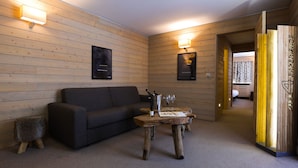A cozy room with a wooden wall, a dark sofa, a wooden coffee table with bottles, and a small stool. - Super G (Courmayeur)