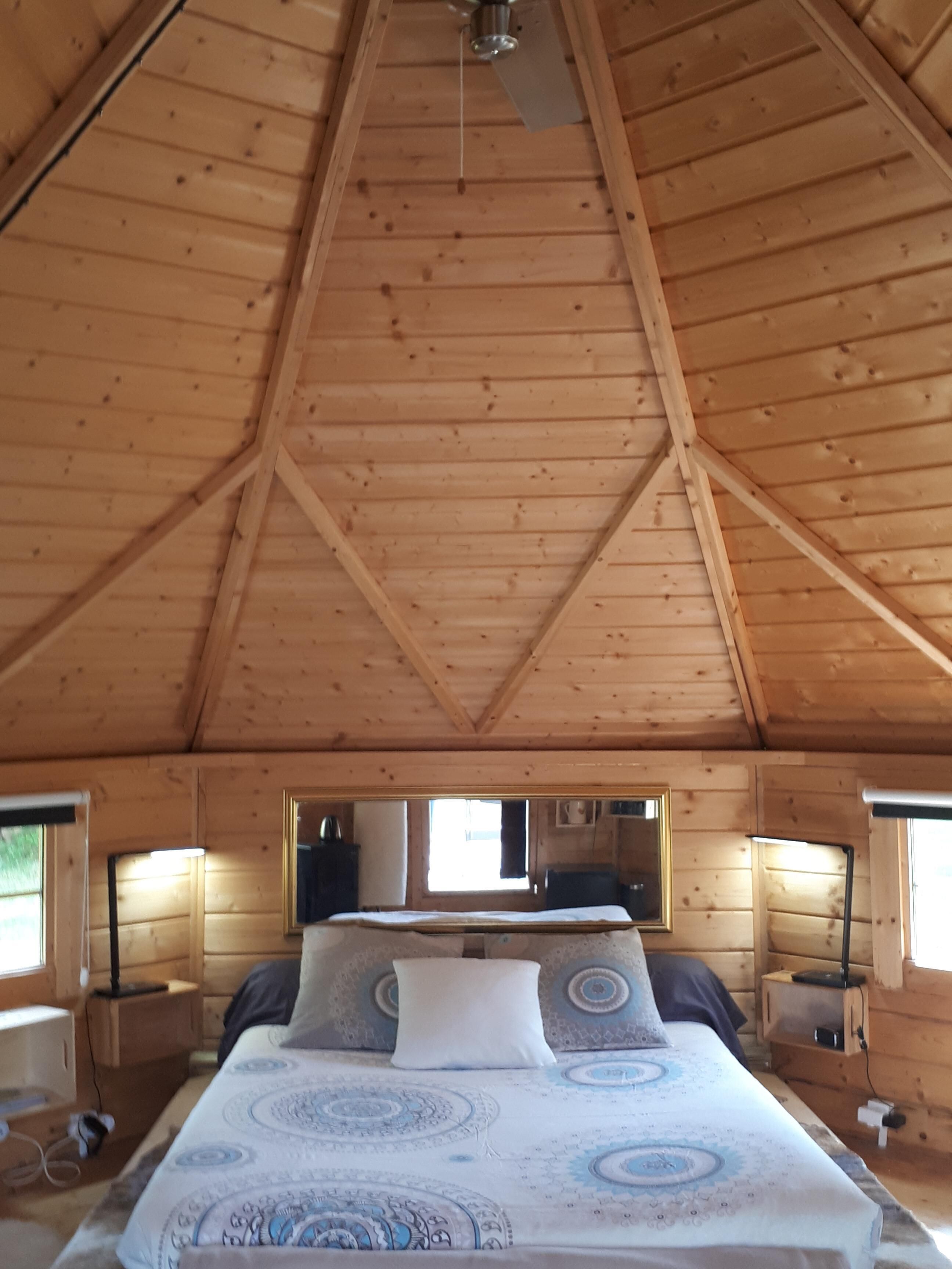 Un dormitorio de cabaña de madera acogedor, con techo alto, cama con ropa de cama estampada, lámparas de noche y ventana con vistas.