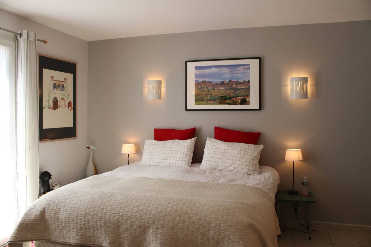 Een netjes opgemaakt bed met een wit-rood geruit beddegoed, twee bedlampen en een ingelijste foto aan de muur.