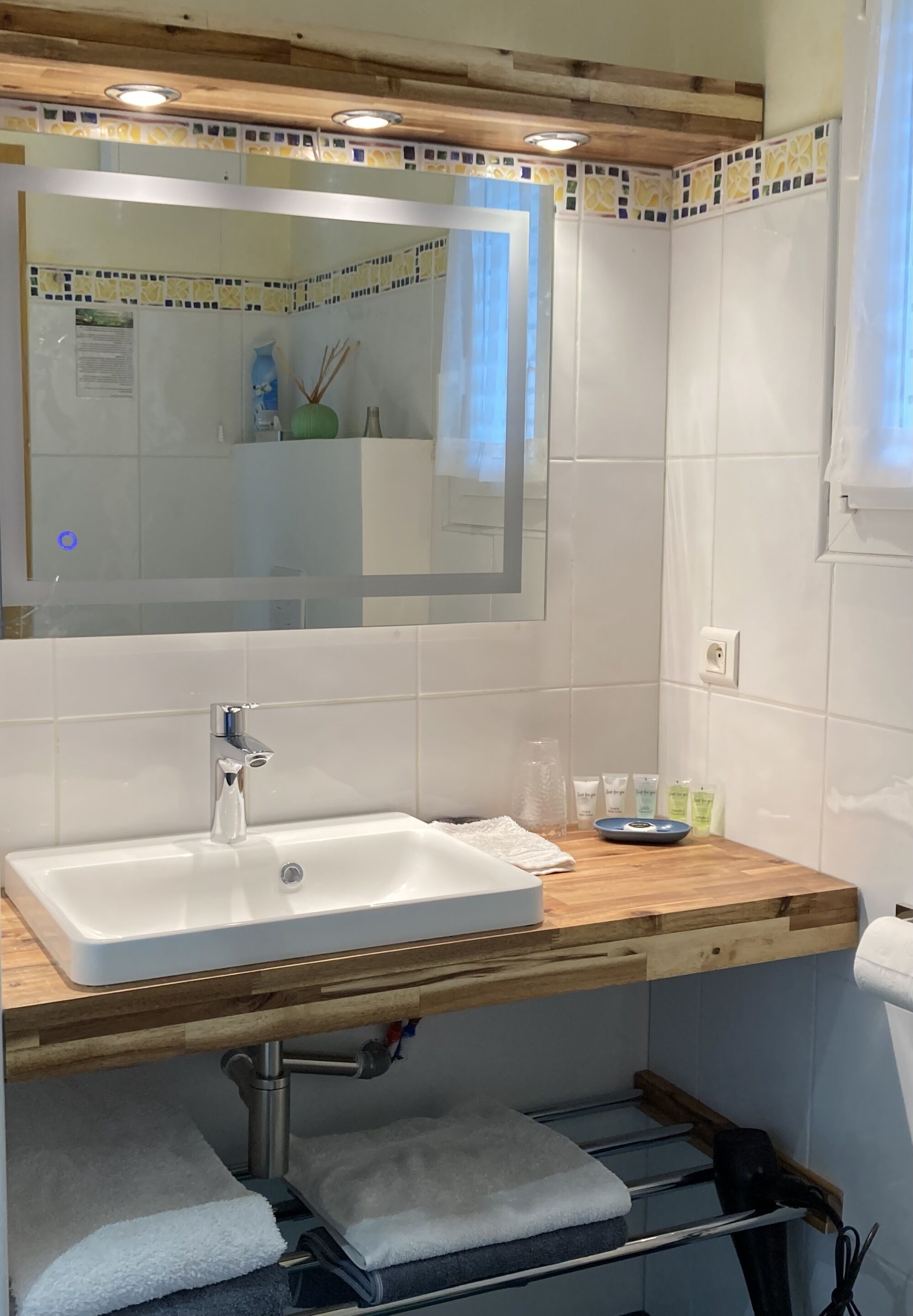 Un baño con un lavamanos blanco, encimera de madera y un espejo grande. Hay artículos de aseo en la encimera y un perchero debajo del lavamanos.