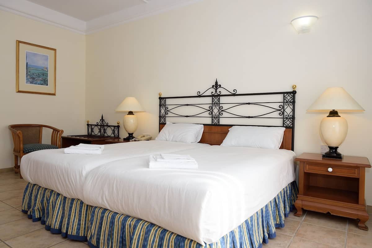 Un lit bien fait, avec du linge de lit blanc et une jupe de lit rayée de bleu et de jaune, deux tables de chevet avec des lampes, une table de nuit en bois, une chaise en osier et un tableau encadré au mur.