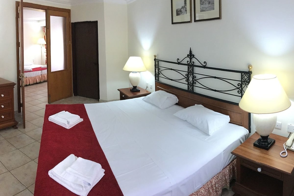 Une chambre d’hôtel avec un lit, deux lampes de chevet, une télévision, une commode en bois, un bureau et une salle de bain visible à travers une porte ouverte.