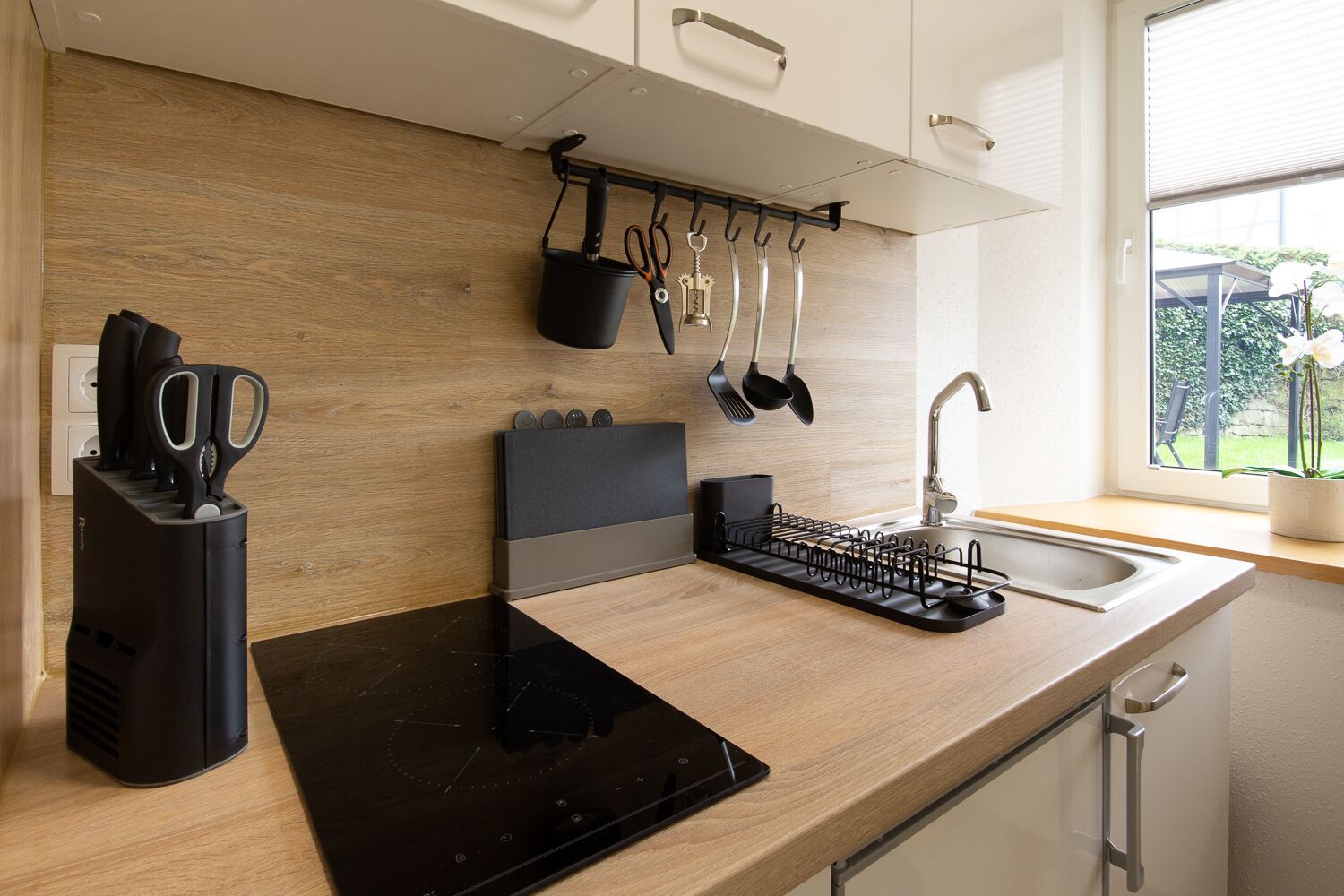 Une cuisine moderne dotée d’une plaque de cuisson à induction noire, d’un bloc à couteaux, de casseroles suspendues et d’une fenêtre donnant sur un paysage verdoyant.